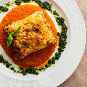 Nahaufnahme einer goldbraun überbackenen Lasagne alla Bolognese, serviert auf fruchtiger Tomatensauce mit Kräuteröl. (Serviervorschlag)