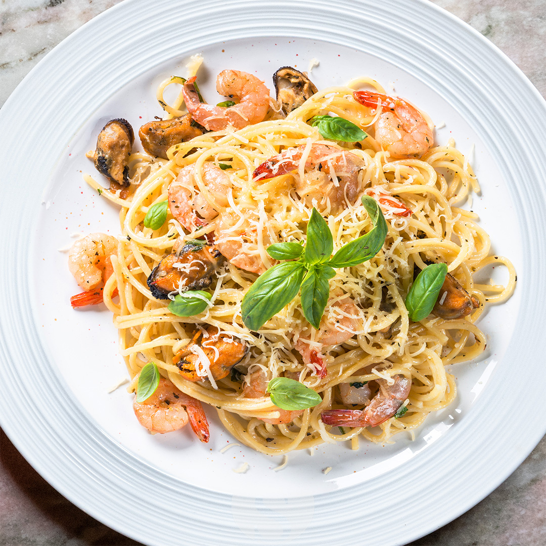 Spaghetti Frutti Di Mare mit Garnelen, Muscheln, Basilikum und geriebenem Käse auf weißem Teller angerichtet (Serviervorschlag)