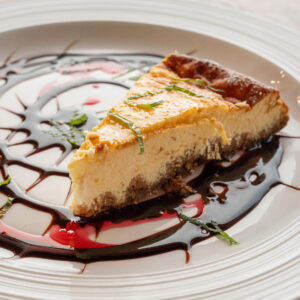 Nahaufnahme eines goldbraun gebackenen Cheesecakes auf einem weißen Teller, dekoriert mit Schokoladen- und Beerensauce sowie frischer Minze. (Serviervorschlag)