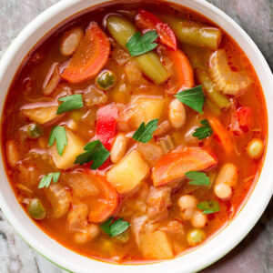 Bunte Minestrone mit Gemüse und Kräutern in weißem Teller auf Marmorplatte (Serviervorschlag)