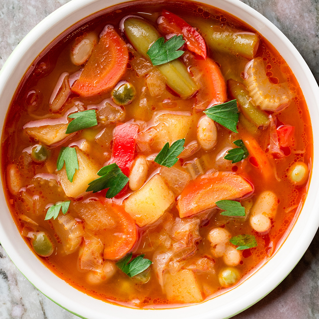 Bunte Minestrone mit Gemüse und Kräutern in weißem Teller auf Marmorplatte (Serviervorschlag)