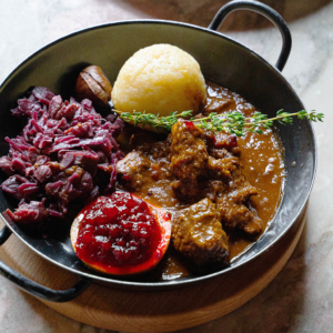 Rehragout mit Erdäpfelknödel, Maroni-Rotkraut und Preiselbeeren in einer schwarzen Pfanne serviert (Serviervorschlag)