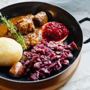 Rehragout mit Erdäpfelknödel, Maroni-Rotkraut und Preiselbeeren in einer schwarzen Pfanne serviert (Serviervorschlag)