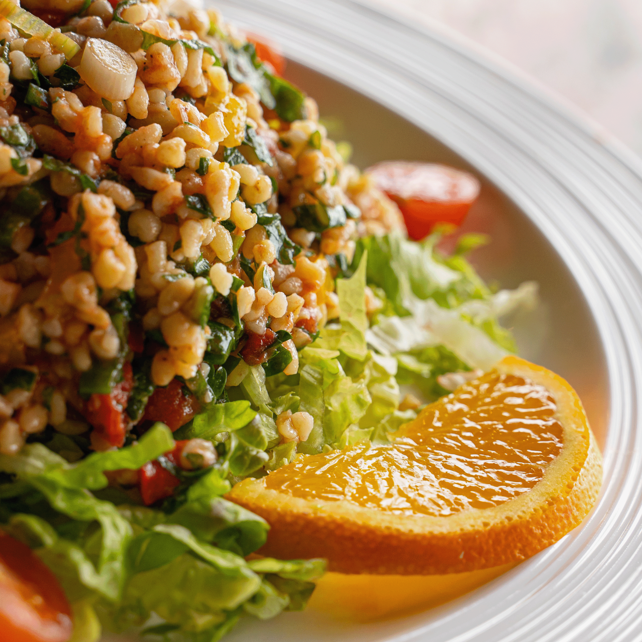 Veganer Bulgursalat mit frischen Kräutern, Blattsalat, Tomaten und Orangenscheiben auf weißem Teller angerichtet (Serviervorschlag)