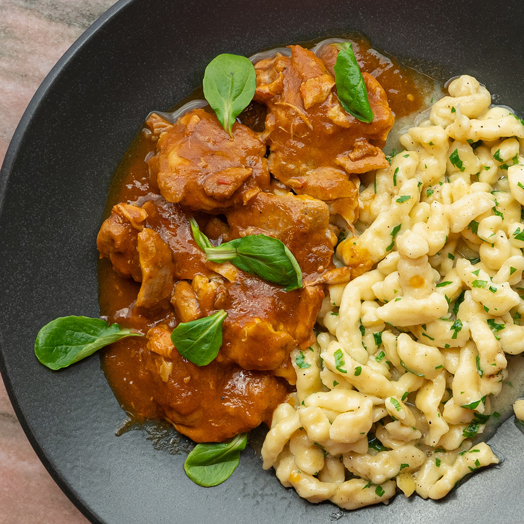 Paprikahendl mit hausgemachten Spätzle in cremiger Paprikasauce, angerichtet auf schwarzem Teller und mit frischen Kräutern garniert. (Serviervorschlag)