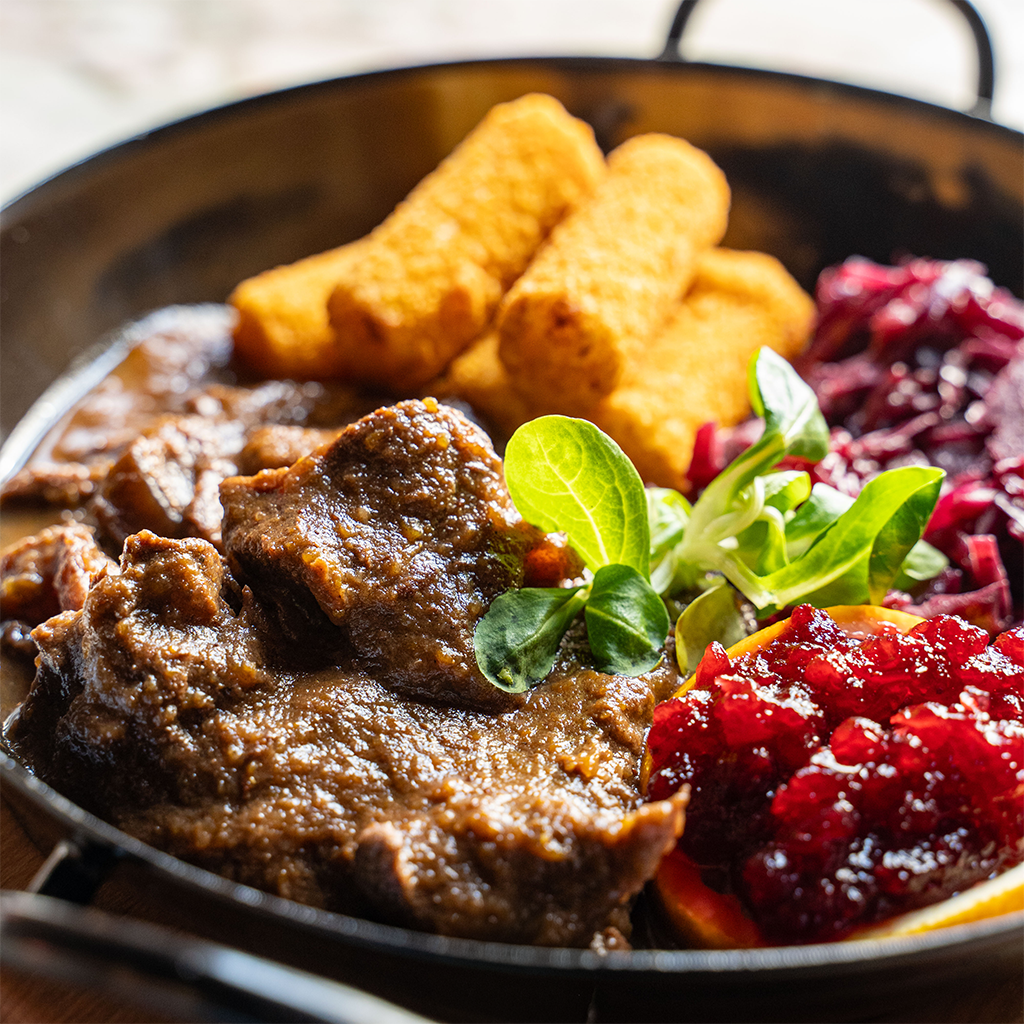 Hirschragout in kräftiger brauner Sauce mit Rotkraut, goldbraunen Kroketten, Preiselbeeren und frischem Blattsalat, serviert in einer dunklen Pfanne. (Serviervorschlag)